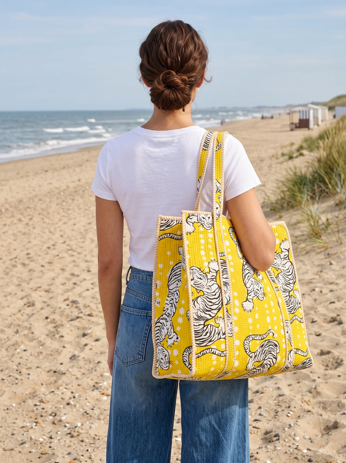 A woman standing on a sandy beach with the ocean in the background, carrying a large yellow canvas tote bag with a black and white tiger print.