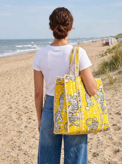 A woman standing on a sandy beach with the ocean in the background, carrying a large yellow canvas tote bag with a black and white tiger print.