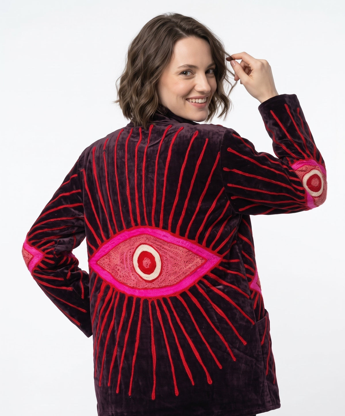 A woman wearing a dark plum velvet jacket with a large embroidered red and pink eye design on the back, shown against a plain white studio background.