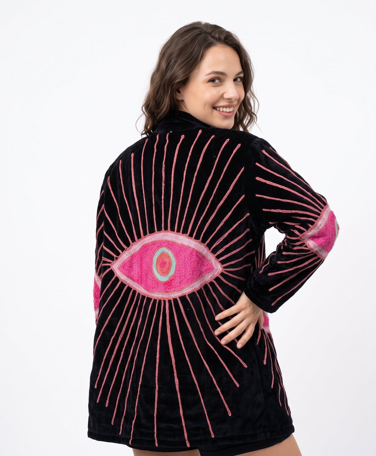A woman modeling a black velvet hooded robe with a large pink and green eye design on the back, accented by pink embroidered rays and pink elbow patches, against a plain white background.