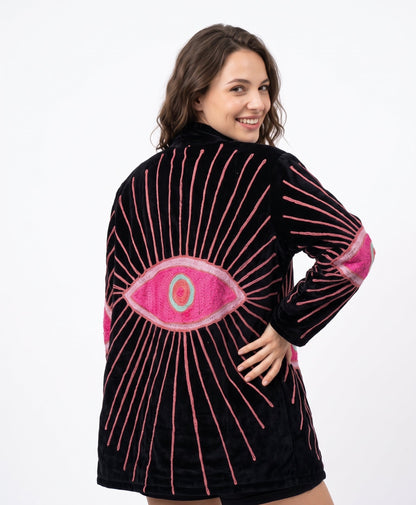 A woman modeling a black velvet hooded robe with a large pink and green eye design on the back, accented by pink embroidered rays and pink elbow patches, against a plain white background.